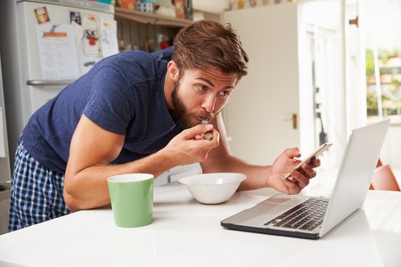 Photo of a young man not eating mindfully. He is eating breakfast while standing and using his mobile phone and laptop.